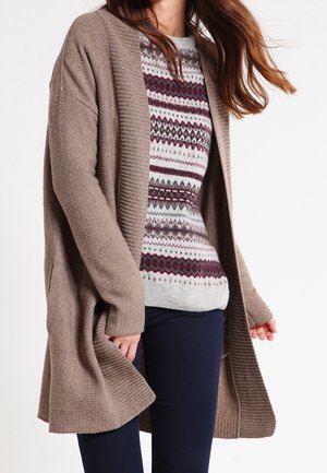 Kobieta ubrana w wzorzysty dziergany sweter w bordowo-szare wzory pod długim, brązowym otwartym kardiganem, w połączeniu z ciemnymi spodniami.