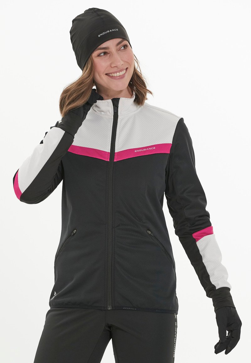 Schwarze und weiße Sportjacke mit pinken Akzenten, mit strukturiertem Obermaterial, Reißverschluss und langen Ärmeln. Kombiniert mit einer schwarzen Mütze.