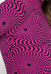 Haut à manches longues ajusté avec un motif en tourbillon noir et rose vif. Le matériau semble texturé, accentuant le design optique.