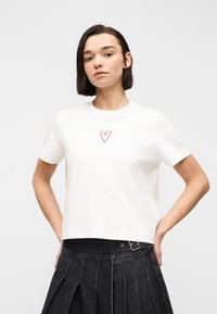 Γυναίκα με κοντά σκούρα μαλλιά, φοράει λευκό μπλουζάκι με μικρή κόκκινη καρδιά και ψηλόμεση μαύρη πλισέ φούστα, με τα χέρια στη μέση.