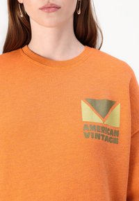 Oranssi collegepaita, valmistettu teksturoidusta kankaasta, jossa on vihreä ja keltainen graafinen logo rinnassa, jossa lukee "AMERICAN VINTAGE".