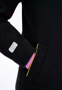 Čierna rukáv s malou nášivkou s nápisom "Lístok do kina, jednorazový vstup" a ruka v vrecku v ružovej rebrovanej rukavici.