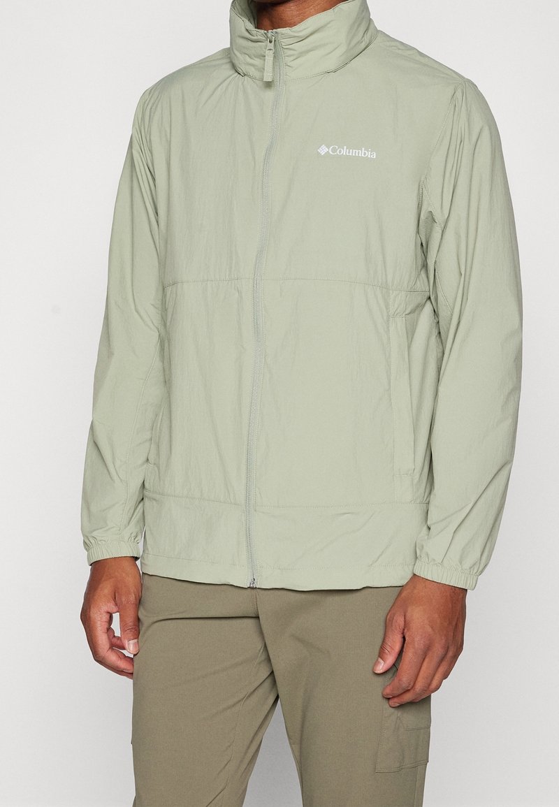 Homme portant une veste zippée Columbia vert clair avec des poignets élastiques et un pantalon vert olive, debout sur un fond uni.
