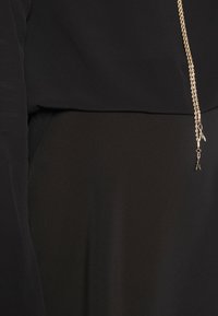 Musta vaate, jossa on sileä ja virtaava kangas, jossa on hienovarainen kiilto. Sisältää metalliketjuelementin, joka on kiinnitetty vyötärölle.