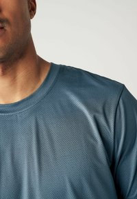 Kék sportpóló strukturált, nedvességet elvezető anyagból. Kerek nyakkivágással és rövid ujjakkal rendelkezik, finoman geometrikus mintázattal.