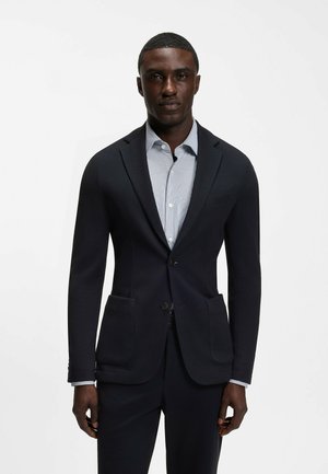 Hombre vistiendo una chaqueta de traje oscuro a medida y pantalones a juego sobre una camisa azul clara abotonada, de pie frente a un fondo blanco liso.
