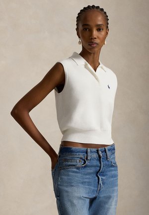 Kvinde med fletninger iført hvid ærmeløs poloshirt og blå jeans, stående med den ene hånd i lommen mod en neutral baggrund.