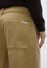 Παντελόνι από βαμβάκι σε χρώμα καμηλό με μια μεγάλη πλαϊνή τσέπη, που διαθέτει κόκκινη και λευκή ετικέτα με το λογότυπο Calvin Klein. Η υφή είναι λεία με κλασική εφαρμογή.