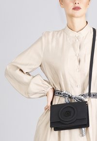 Černá crossbody taška vyrobená z hladkého materiálu, s kulatým embosovaným logem, sklápěcí klopou a odnímatelným popruhem.