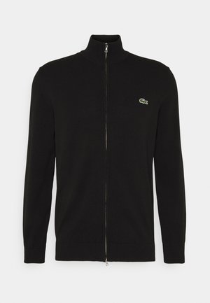 Schwarzer Zip-Pullover mit hohem Kragen, aus Strickstoff gefertigt. Mit gerippten Bündchen und Saum, versehen mit einem kleinen grünen Logo auf der linken Brust.