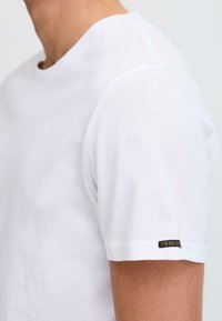 Camiseta de algodón blanca con mangas cortas, cuello redondo y una pequeña etiqueta negra en la manga. Textura suave, corte estándar y diseño minimalista.