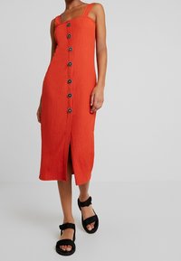 Punainen pellavamekko midimittaisena leveillä olkaimilla, etunapeilla ja etuhalkiolla. Yhdistettynä mustiin sandaaleihin, joissa on paksut remmit ja tasapohja.