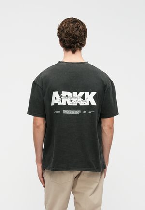 T-shirt de algodão cinza escuro com um corte boxy, apresentando gráficos e texto de logotipo brancos nas costas. Mangas curtas e decote redondo.