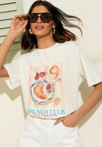 Valkoinen puuvillapaita, jossa on värikäs brunssiteemainen grafiikka, joka sisältää croissantin, kahvin, mansikoita ja "BRUNCH CLUB" -tekstiä sinisessä ja punaisessa.
