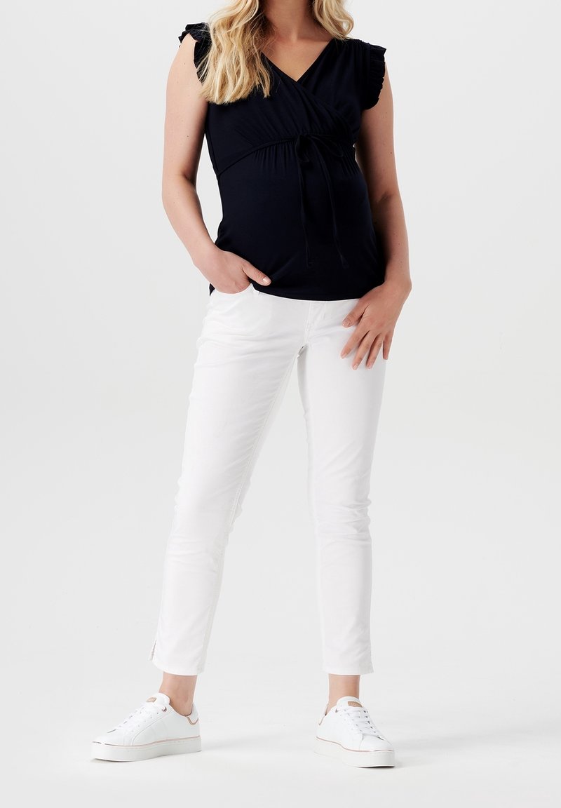 Marineblaues Wickeloberteil mit Rüschensäumen, kombiniert mit schmalen weißen Hosen und weißen Sneakers. Besitzt eine Krawattendetails an der Taille und seitliche Schlitze an den Manschetten.