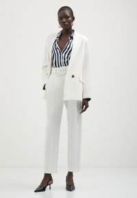 Weißer maßgeschneiderter Anzug mit weitem Beinkleid und Blazer, kombiniert mit einem blau-weißen gestreiften Hemd. Schwarze spitze Schuhe vervollständigen den Look.