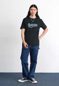 Črna bombažna majica s svetlo modrim logotipom "Converse", v kombinaciji z modsko kavbojkami in črnimi supergami na teksturiranem tleh proti enotnemu ozadju.