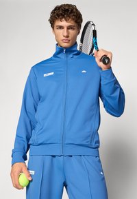 Niebieska sportowa kurtka i spodnie z gładkiego materiału, z wysokim kołnierzem, zapięciem na zamek i akcentami z logo. Model trzyma rakietę do tenisa i piłkę.