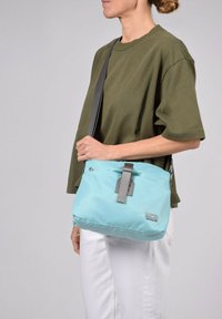 Sac à épaule bleu clair en matériau durable, avec une sangle grise et des accents en métal, orné d'un écusson de logo et d'une texture lisse.