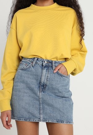 Femme portant un top jaune ample à manches longues, rentré dans une mini-jupe en denim bleu clair taille haute, une main dans la poche.
