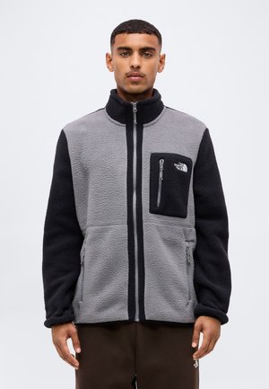 Ein Mann steht frontseitig und trägt eine grau-schwarze Fleecejacke mit Reißverschlusstaschen und dem The North Face-Logo auf der Brusttasche.