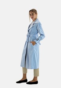 Ljusblå trenchcoat med skärp i midjan, långa ärmar och kontrasterande krage. Modellen bär beige byxor och svarta ballerinaskor. Slät textur.