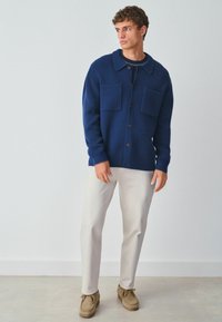 Granatowy dzianinowy kardigan z kołnierzem, zapięcie na guziki z przodu, dwie duże kieszenie naszywane, w połączeniu z jasnobeżowymi spodniami i brązowymi zamszowymi butami.