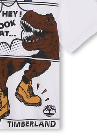 Bela majica s kratkimi rokavi, ki prikazuje rjav dinozaver grafiko, oblečenega v rumene škornje. Črn napis in detajli z logotipom Timberland na dnu.