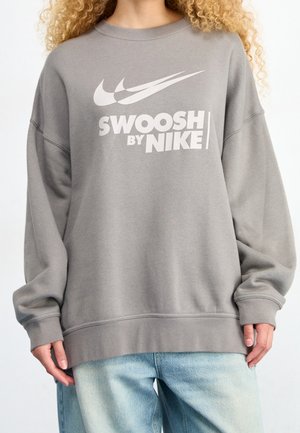 Person iført en løs grå sweatshirt med hvid "Swoosh by Nike" tekst og logoer, kombineret med lyseblå denimjeans.