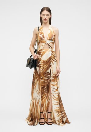 Woman wearing a floor-length brown and cream tropical print dress with cutouts, holding a black handbag with a gold chain strap.