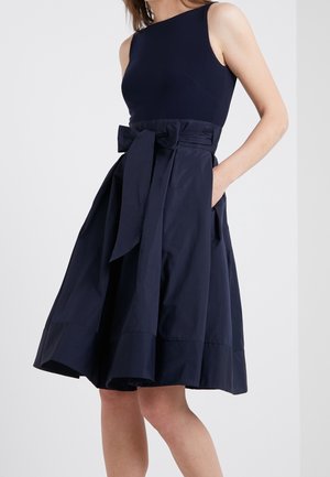 Woman wearing a sleeveless navy dress with a fitted bodice, flared skirt, large bow at waist, and hands in pockets against a plain background.