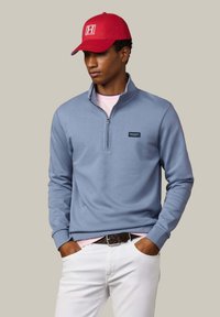 Jonge man met een rode pet, lichtblauwe trui met een kwartrits, lichtroze overhemd, witte broek en een donkerbruine riem, handen in de zakken.