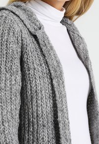 Szary dzianinowy kardigan o fakturowanym wykończeniu, z otwartym przodem i subtelnym kapturem, noszony na białym golfie. Miękki, przytulny materiał z wełny.