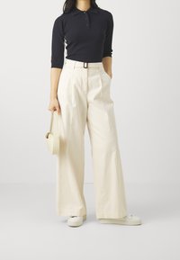 Zwarte polo met drie knopen, gecombineerd met hoge crèmekleurige wijde broek en witte sneakers, terwijl een kleine beige tas wordt vastgehouden.