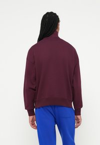 Bordeaux pullover sweatshirt med høj krave, drop shoulders og ribbede manchetter, parret med blå bukser. Glat tekstur, afslappet pasform.