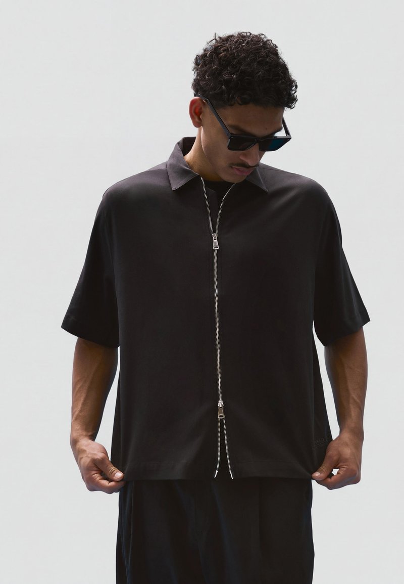 Homme portant une chemise noire à manches courtes avec une double fermeture éclair verticale, lunettes de soleil foncées, regardant vers le bas, tenant les côtés de la chemise sur un fond uni.