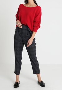 Röd överdimensionerad stickad tröja, högmidjade svart och grå rutig byxor med bälte, och svarta loafers. Slät textur och avslappnad passform.