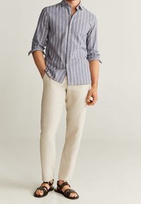 Homme portant une chemise à rayures verticales bleues et blanches, un pantalon beige, et des sandales à bouts ouverts marron foncé, debout devant un fond uni.