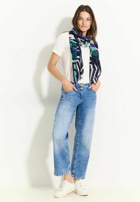 Lyseblå denim jeans med en afslappet pasform, parret med en hvid t-shirt og et mønstret tørklæde i blå, grøn og lilla, afsluttet med hvide sneakers.