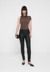 Haut asymétrique marron associé à un legging noir ajusté et des escarpins noirs à bout pointu. Une petite pochette noire est tenue dans la main gauche.