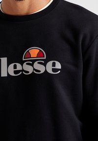 Sweatshirt noir arborant un grand logo avec le mot "lesse" en gris, agrémenté d'un emblème coloré ressemblant à un soleil en orange et rouge.