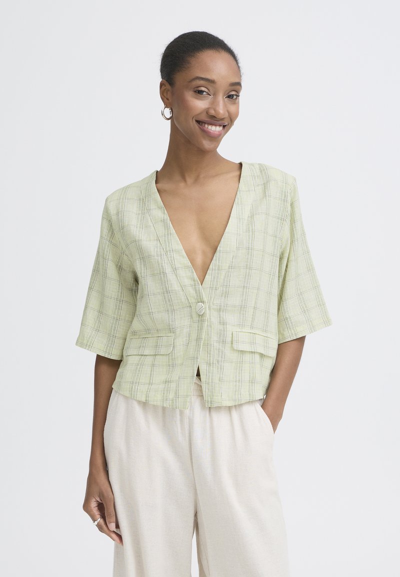 Femme souriante, portant un blazer à manches courtes à carreaux verts clairs avec un col en V profond et un pantalon crème à jambes larges, debout devant un fond blanc.