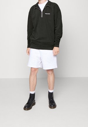 Felpa nera con zip e logo "PEGALDOR", abbinata a pantaloni corti grigi chiari. Stivali neri con lacci, calze bianche, in piedi contro uno sfondo neutro.