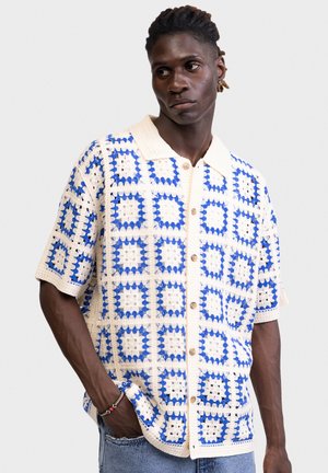 Mężczyzna w niebiesko-białej szydełkowanej koszuli z krótkim rękawem na guziki z kołnierzykiem, stojący z jedną ręką w kieszeni na jednolitym tle.