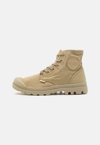 Botas de tobillo de lona color beige con puntera de goma, suela texturizada y ojales de metal, con un parche circular de logo en el lado exterior.