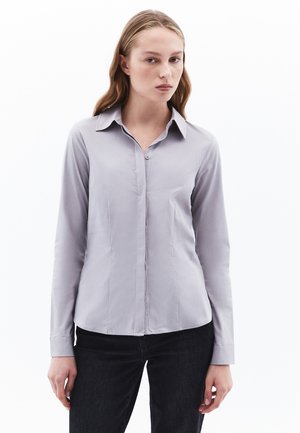 Camisa de manga larga gris claro con botón en el frente, cuello puntiagudo, ajuste slim y textura suave, combinada con jeans oscuros.