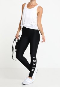 Witte mouwloze tanktop met een knoop in de taille, gecombineerd met zwarte leggings met een witte "NIKE"-logo langs het been. Witte sportschoenen.