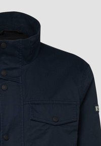 Tumši zils žakete ar augstu apkakli, pogātu krūšu kabatu ar pārsegu, priekšējām sprādzēm un logotipa plāksteri uz augšējā kreisā piedurknes.