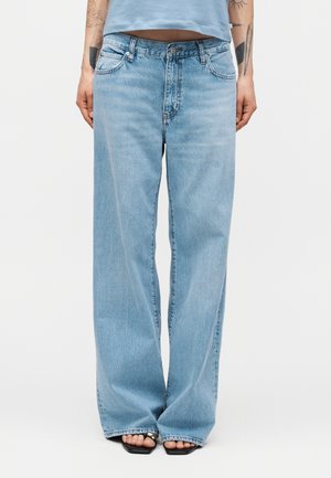 Osoba ubrana w jasnoniebieskie dżinsy o szerokich nogawkach i czarne sandały z odkrytymi palcami, z widocznymi tatuażami na przedramionach, stojąca na jednolitym tle.