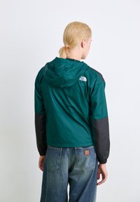 Groene en zwarte windbreaker jas met een capuchon, voorzien van een logo op de achterkant, gemaakt van een lichtgewicht materiaal. Aansluitende mouwen met elastische boorden.